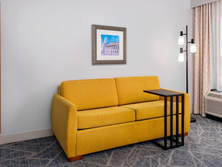Quarto de hotel com sofá amarelo, um quadro na parede, abajur e janela com cortinas.
