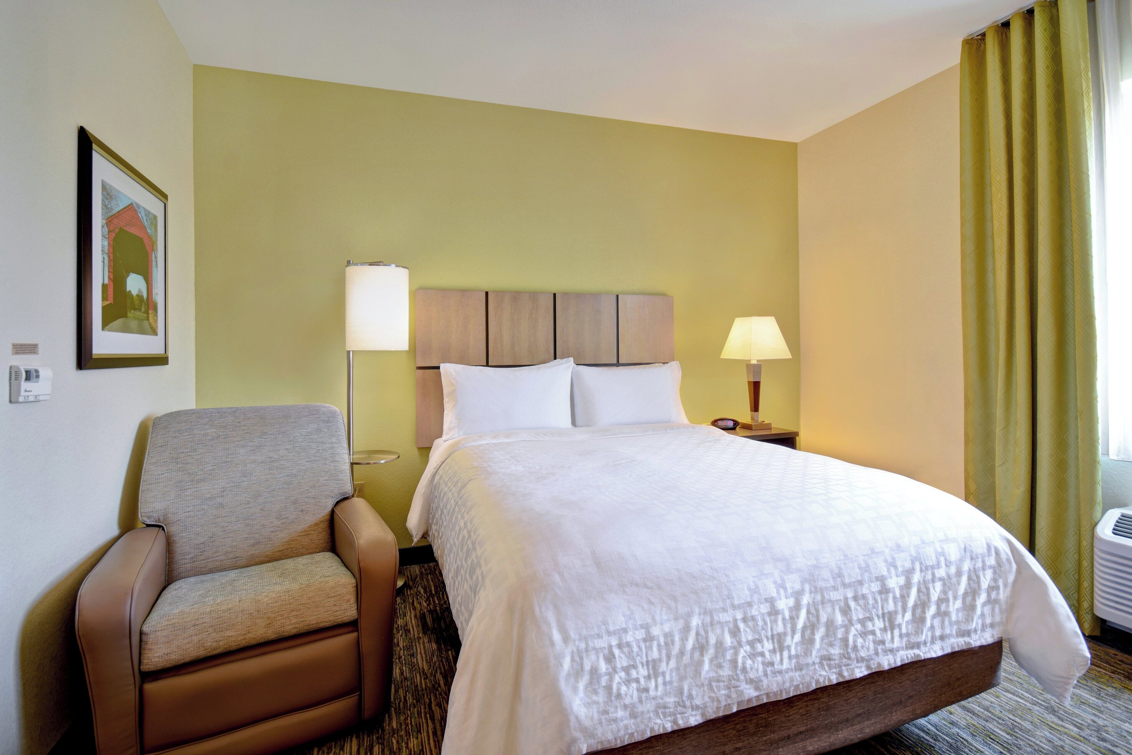 A hotel room with a bed, a chair, two bedside lamps, a framed picture on the wall, and a window with curtains.