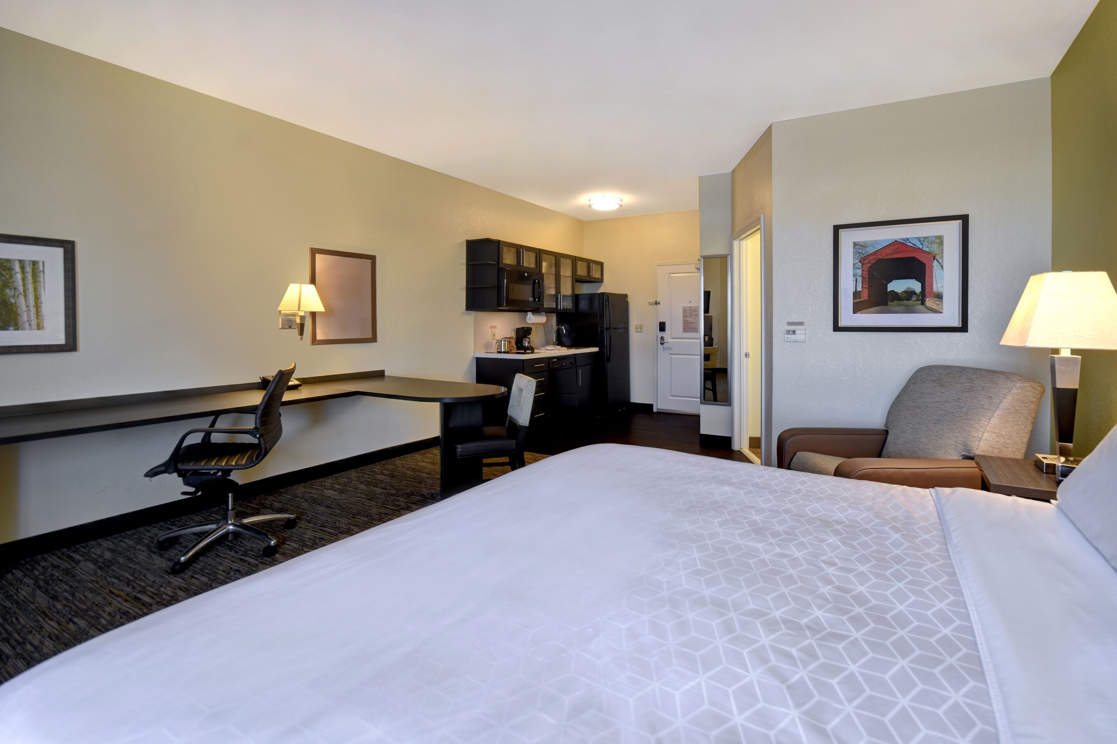A hotel room with a bed, a desk, a kitchenette, and a chair.