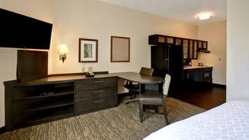 Habitación de hotel con cama, escritorio con silla, televisor de pantalla plana, lámpara, cuadros enmarcados y kitchenette.