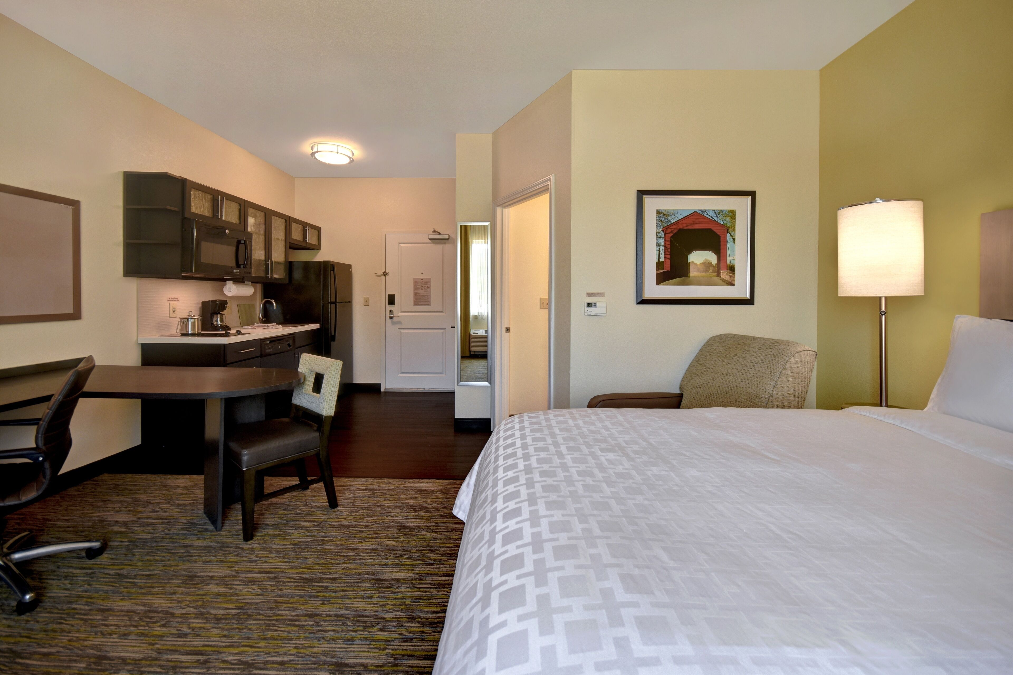 Quarto de hotel com cama, escrivaninha, cadeira, bancada de cozinha e um quadro na parede.