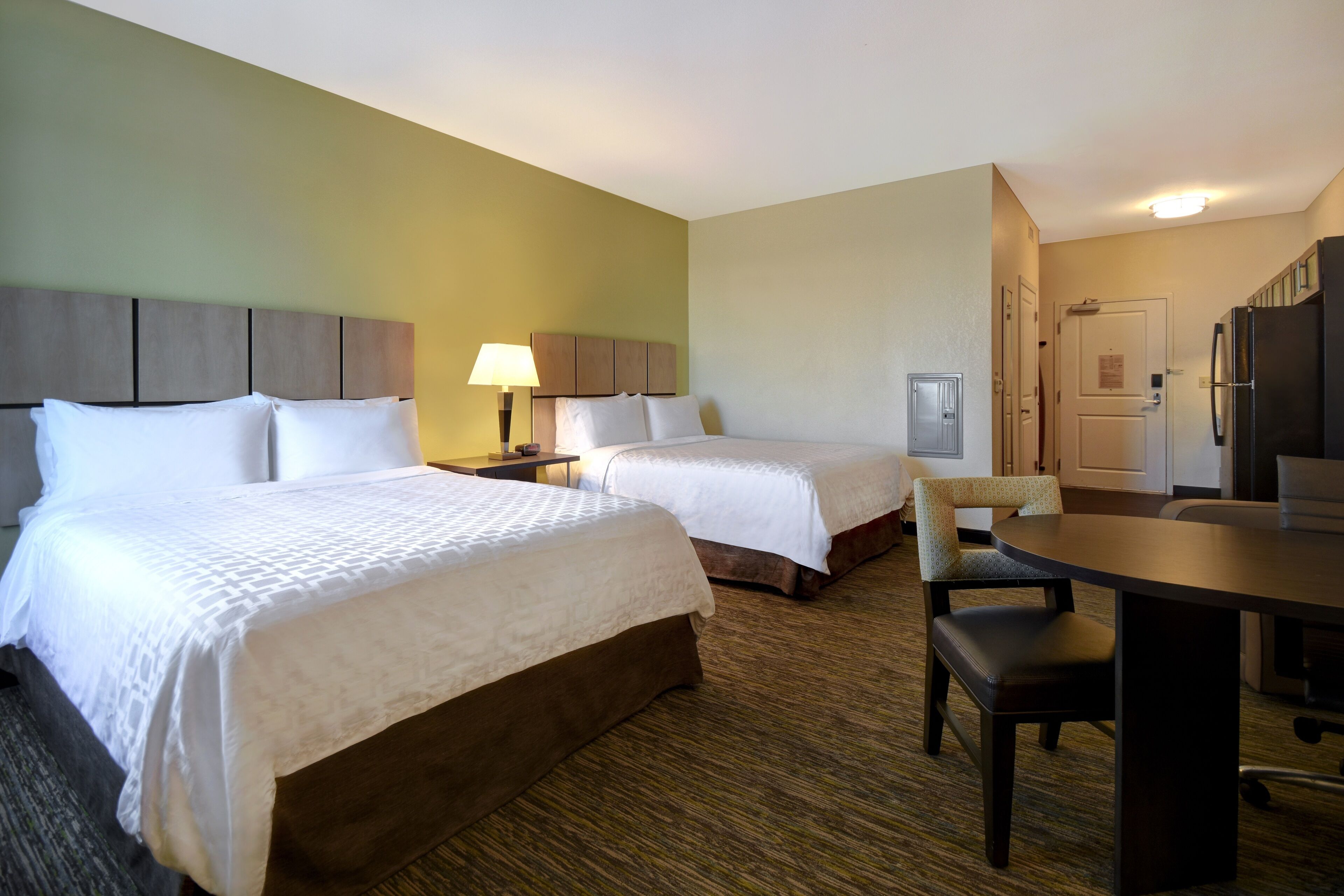A hotel room with two beds, a desk, a chair, a lamp, a refrigerator, and a door.