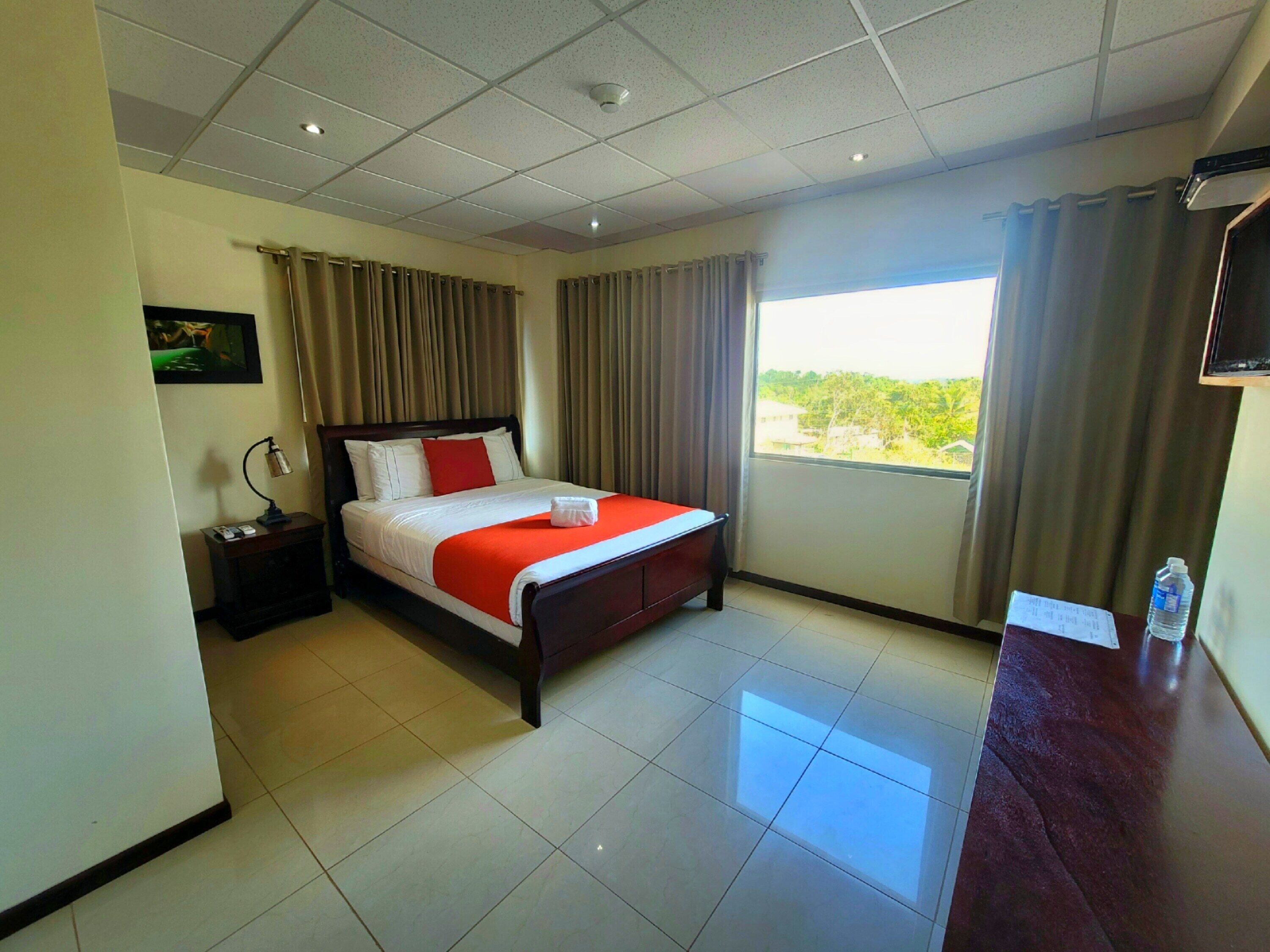 A hotel room with a bed, a nightstand, a desk, a chair, a TV, and a window with curtains.