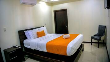A neatly made bed with white and orange bedding, a dark wooden headboard, a small bedside table, and a chair to the right.