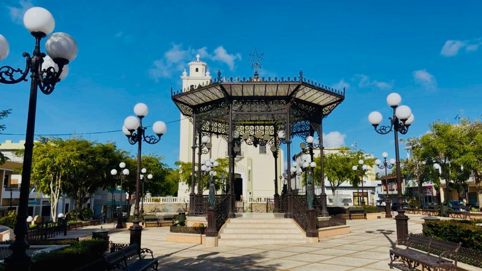 A gazebo with ornate metalwork and a star on top, surrounded by lamp posts and benches in a plaza with a clear sky.