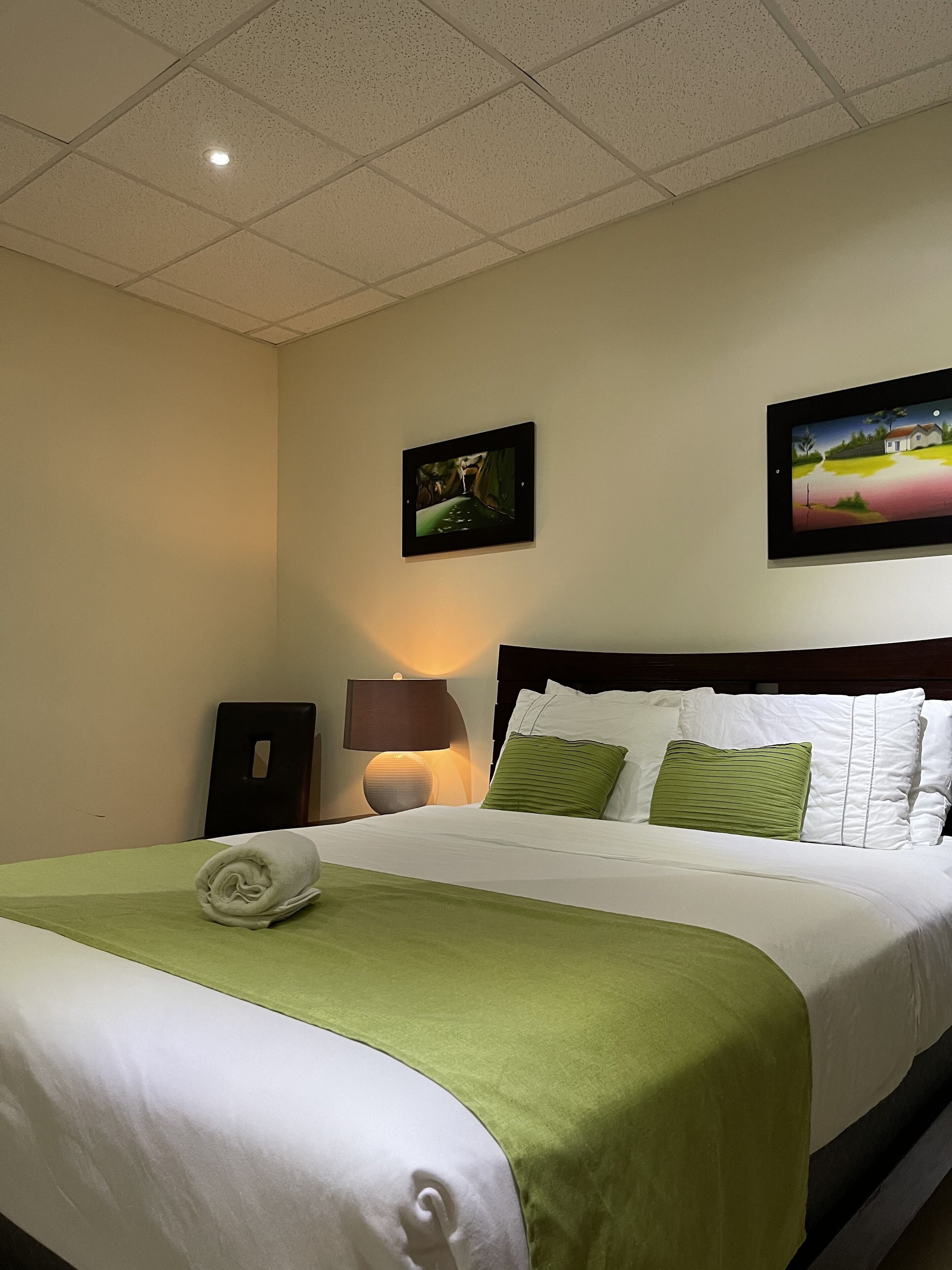 A hotel room with a neatly made bed, a nightstand with a lamp, and two framed pictures on the wall.