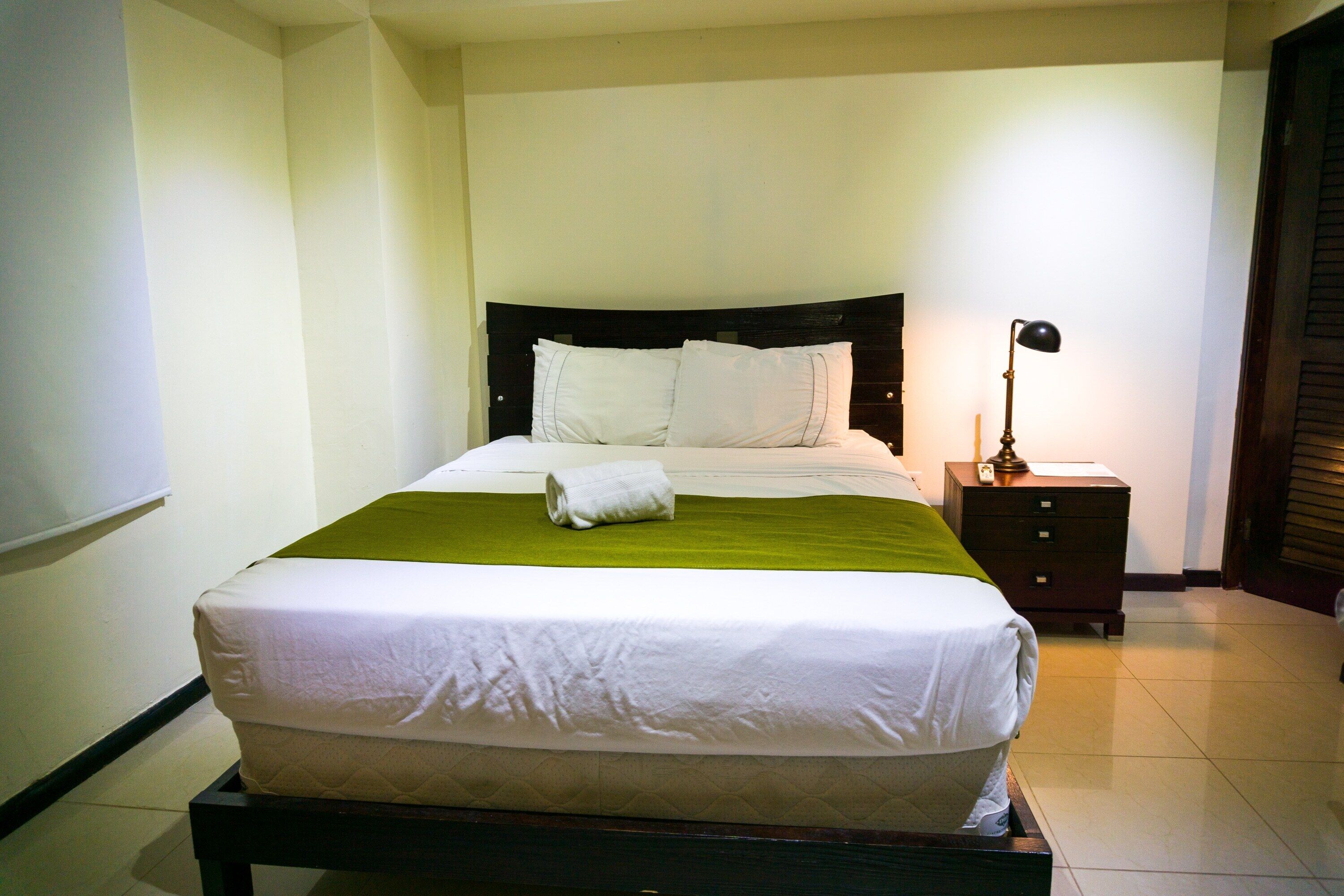 A neatly made bed with a green bedspread and white linens, a bedside lamp, and a wooden nightstand.