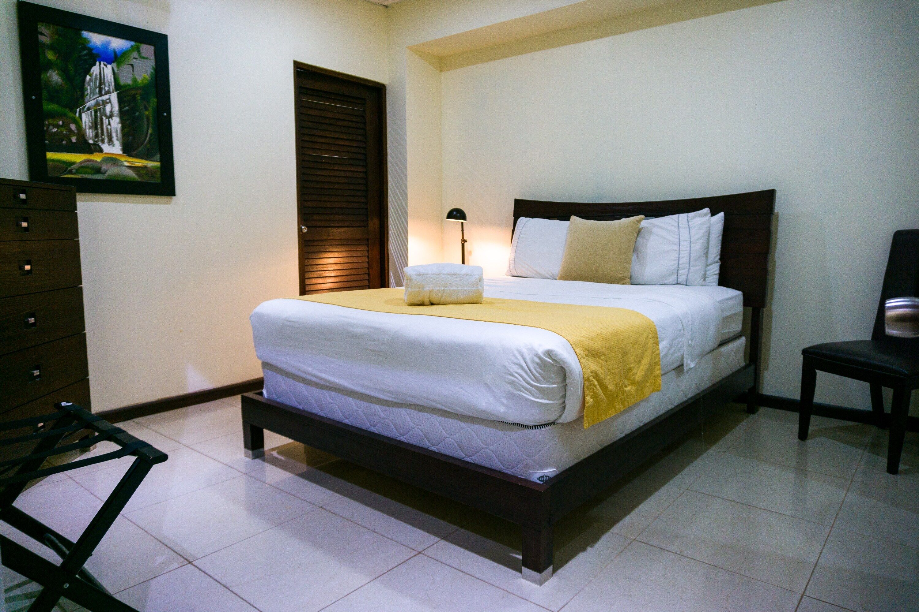 Una cama bien hecha con un edredón amarillo, una lámpara de noche y un cuadro en la pared.