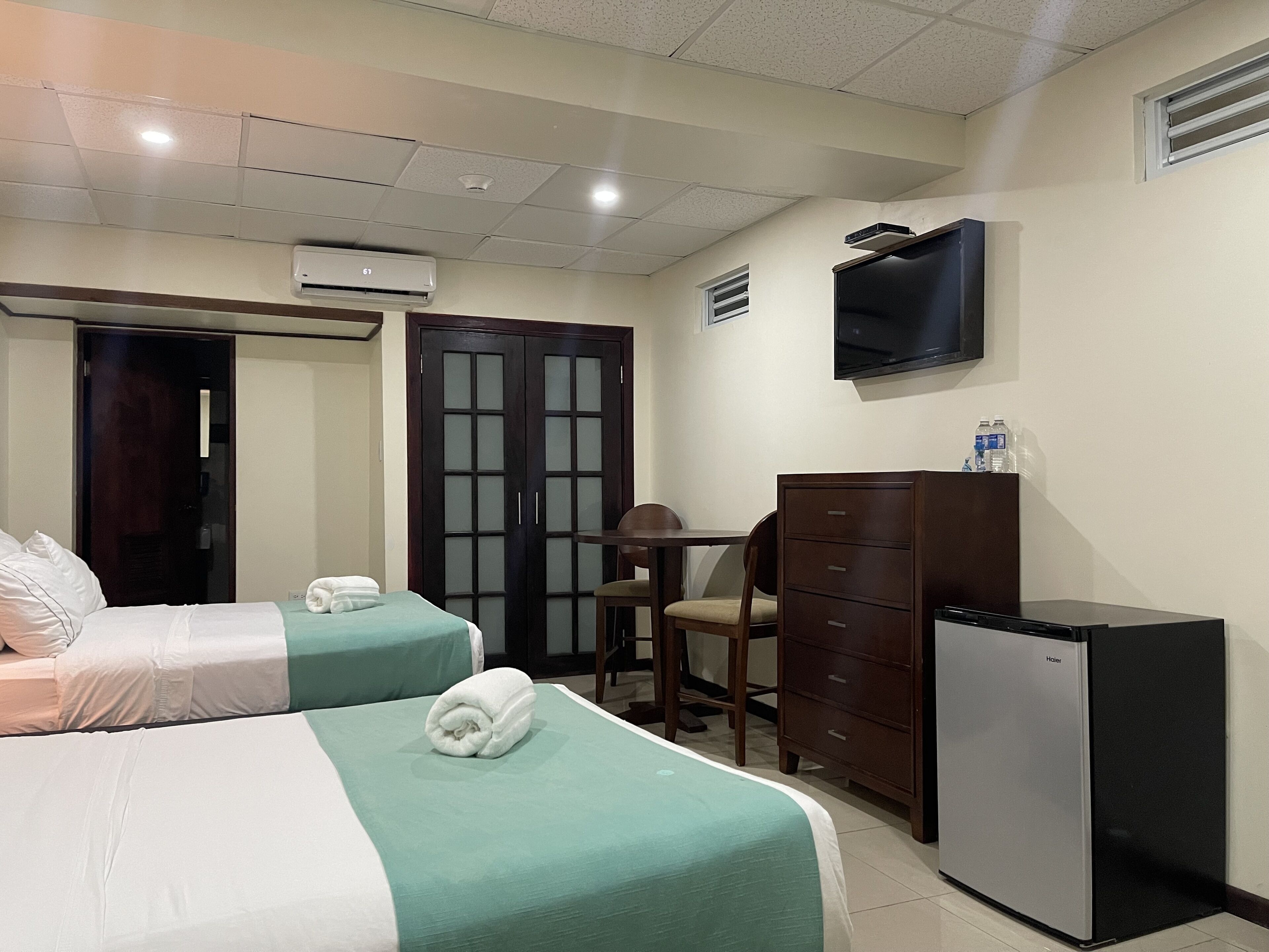 A hotel room with two beds, a desk, a chair, a dresser, a television, and an air conditioning unit.