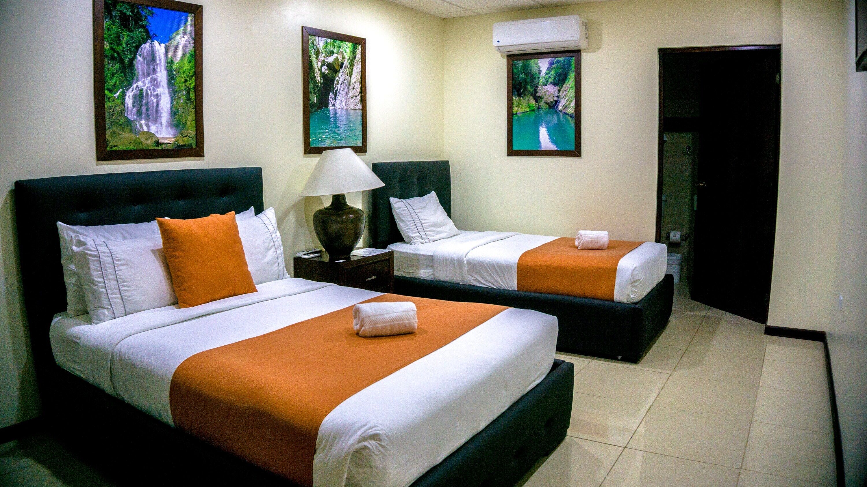 Una habitación de hotel con dos camas, cada una con ropa de cama blanca y naranja, una mesita de noche con lámpara y cuadros en las paredes.