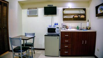 A compact kitchenette with a microwave, coffee maker, and small refrigerator. There is a chair and a table, and a shelf with dishes.