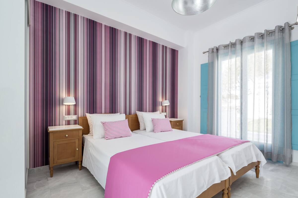 A bedroom with a bed, two bedside tables, a nightstand, a window with curtains, and a striped wall.