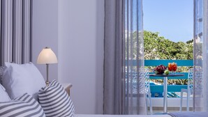 A hotel room with a bed, a bedside lamp, a table with drinks and fruit, and a view of greenery outside. - Santellini Hotel (Santorini)