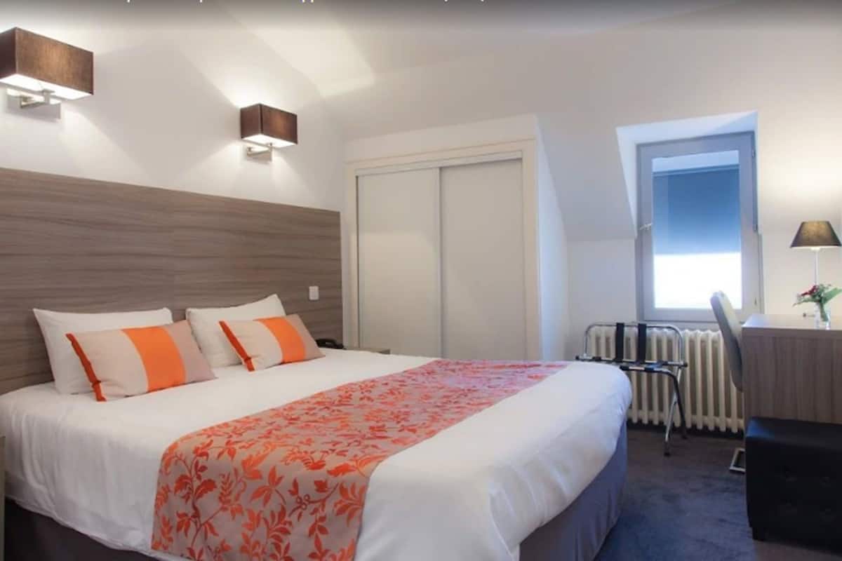 Una habitació d'hotel amb un llit, una taula, una cadira, un radiador i una finestra.