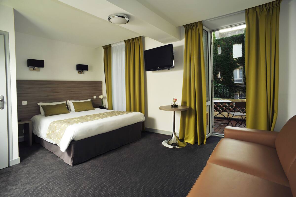Habitació d'hotel amb llit, taula petita, televisió i balcó amb cortines grogues.