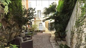 A narrow courtyard with a gravel path, surrounded by potted plants and a seating area with chairs.