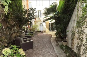 A narrow courtyard with a gravel path, surrounded by potted plants and a seating area with chairs.