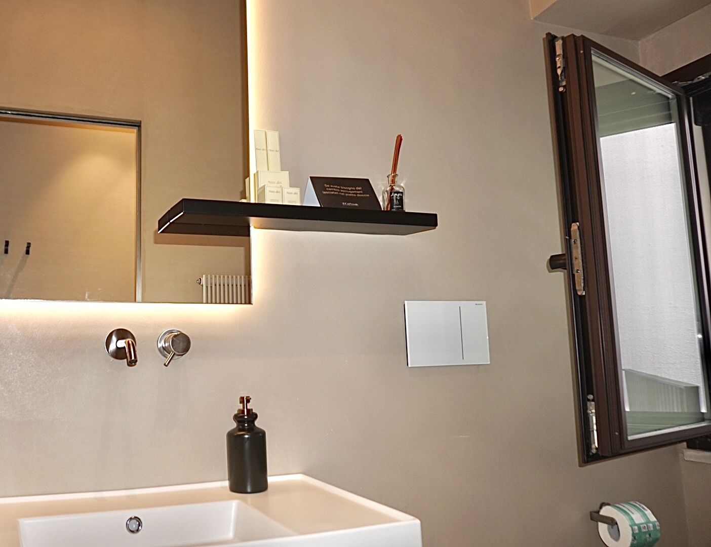 Un baño con lavamanos blanco, un espejo, una repisa con artículos de aseo y una ventana con vistas.