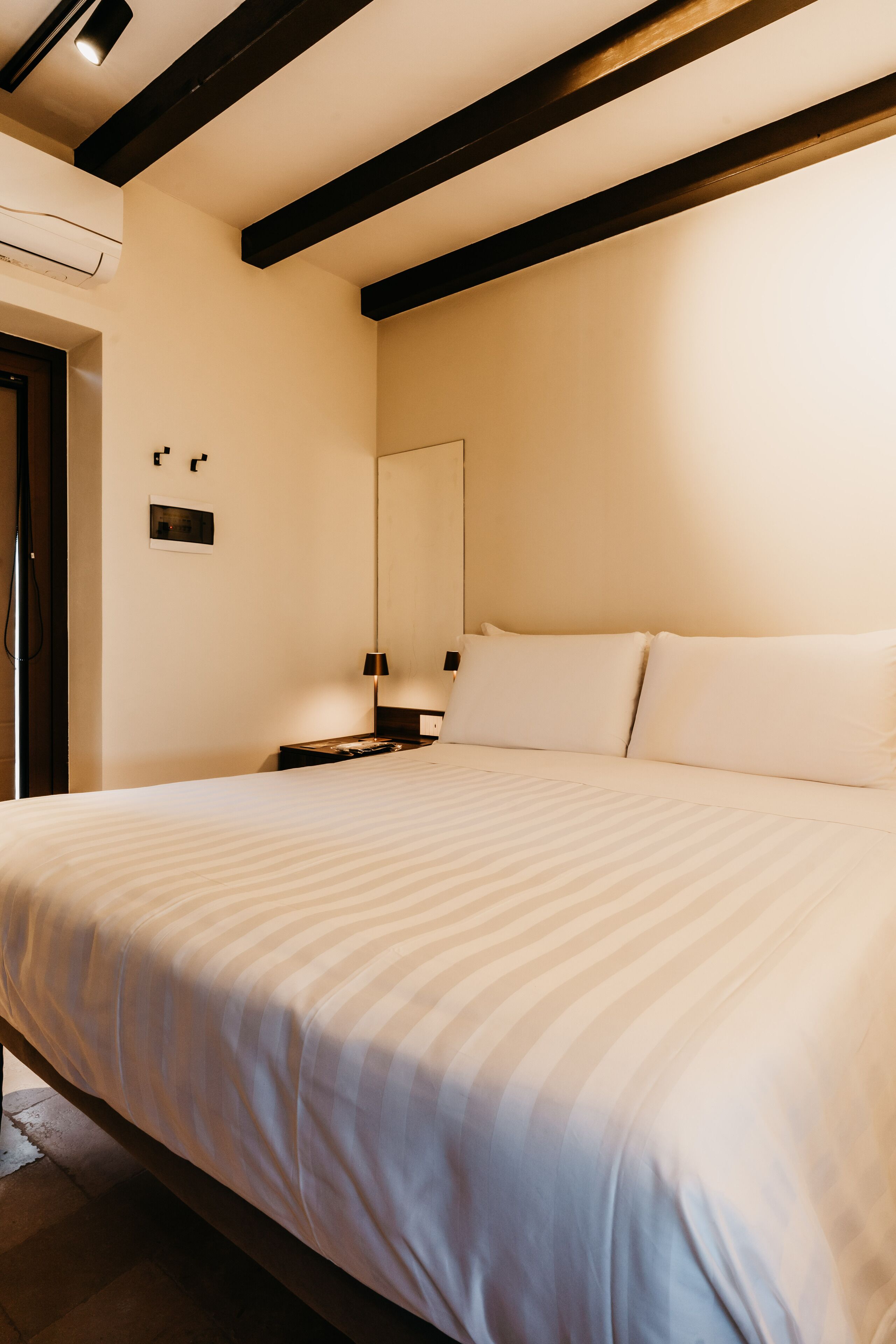A hotel room with a large bed, a bedside table, a mirror, and a wall-mounted air conditioning unit.