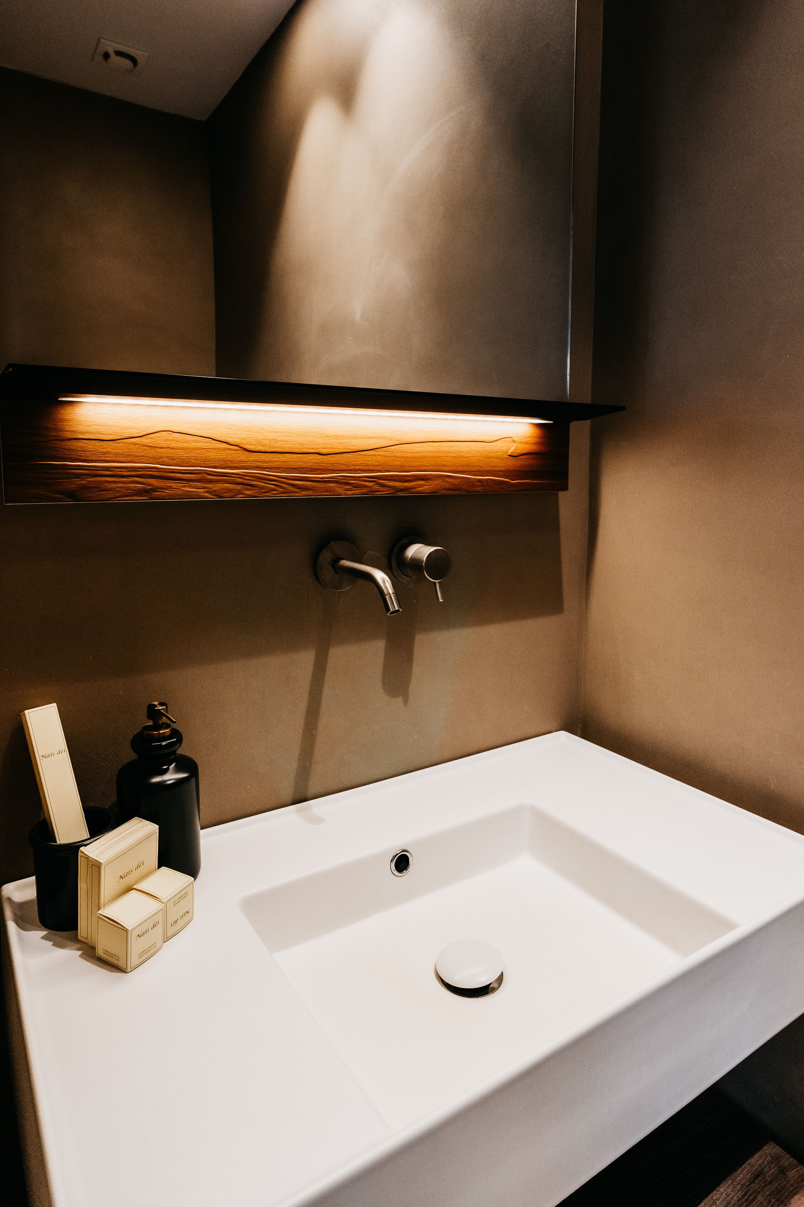 Un baño moderno con un lavamanos blanco, un estante de madera encima y un aplique en la pared.