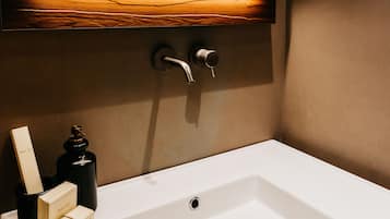 Un baño moderno con un lavamanos blanco, un estante de madera encima y un aplique en la pared.