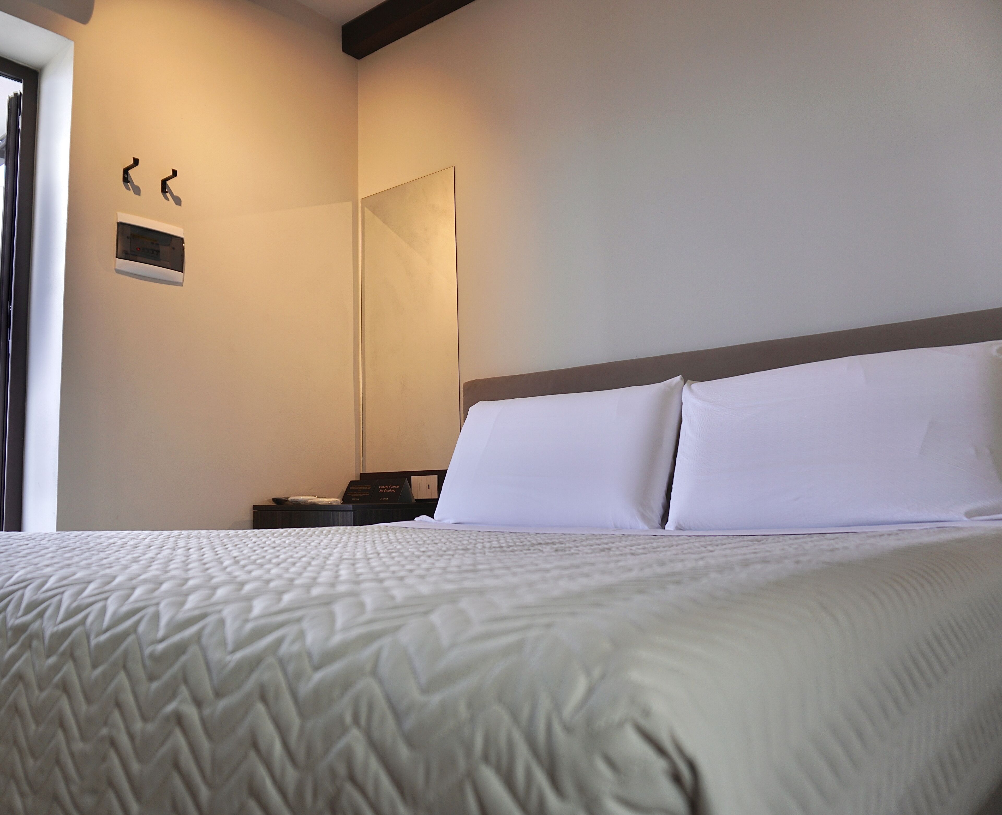 A neatly made bed with a textured blanket, a bedside table with a lamp, and a wall-mounted thermostat.