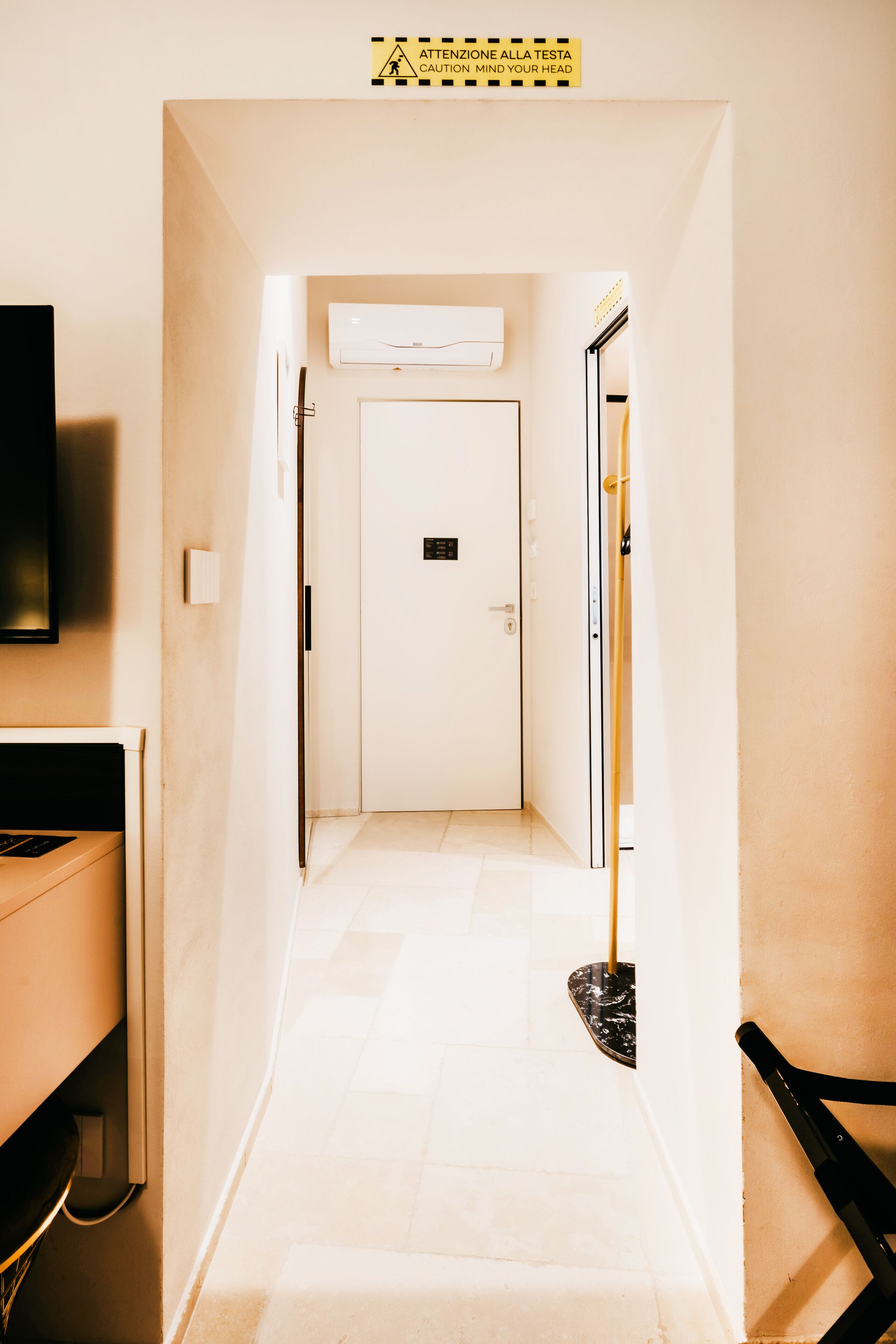 A narrow hallway with a white door at the end, a sign above warning to mind your head, and a black chair on the right.