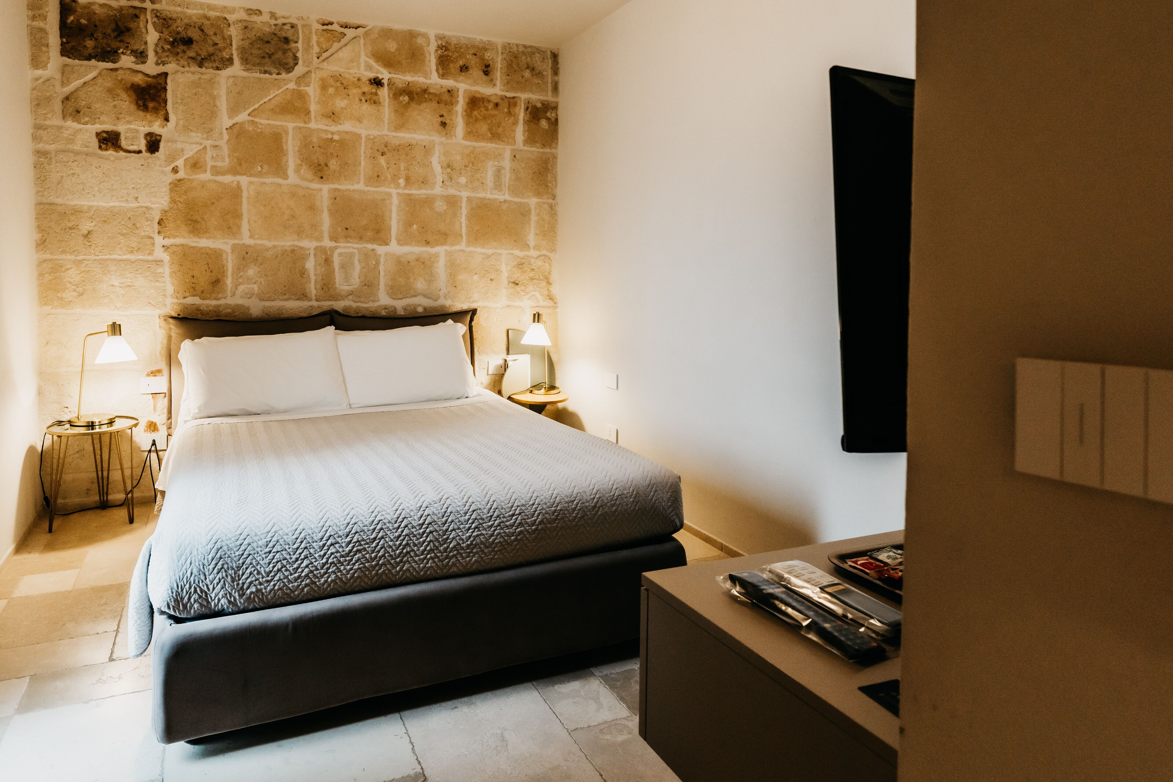 Quarto com parede de pedra, cama com roupa de cama branca, duas luminárias de cabeceira e uma TV de tela plana na parede.