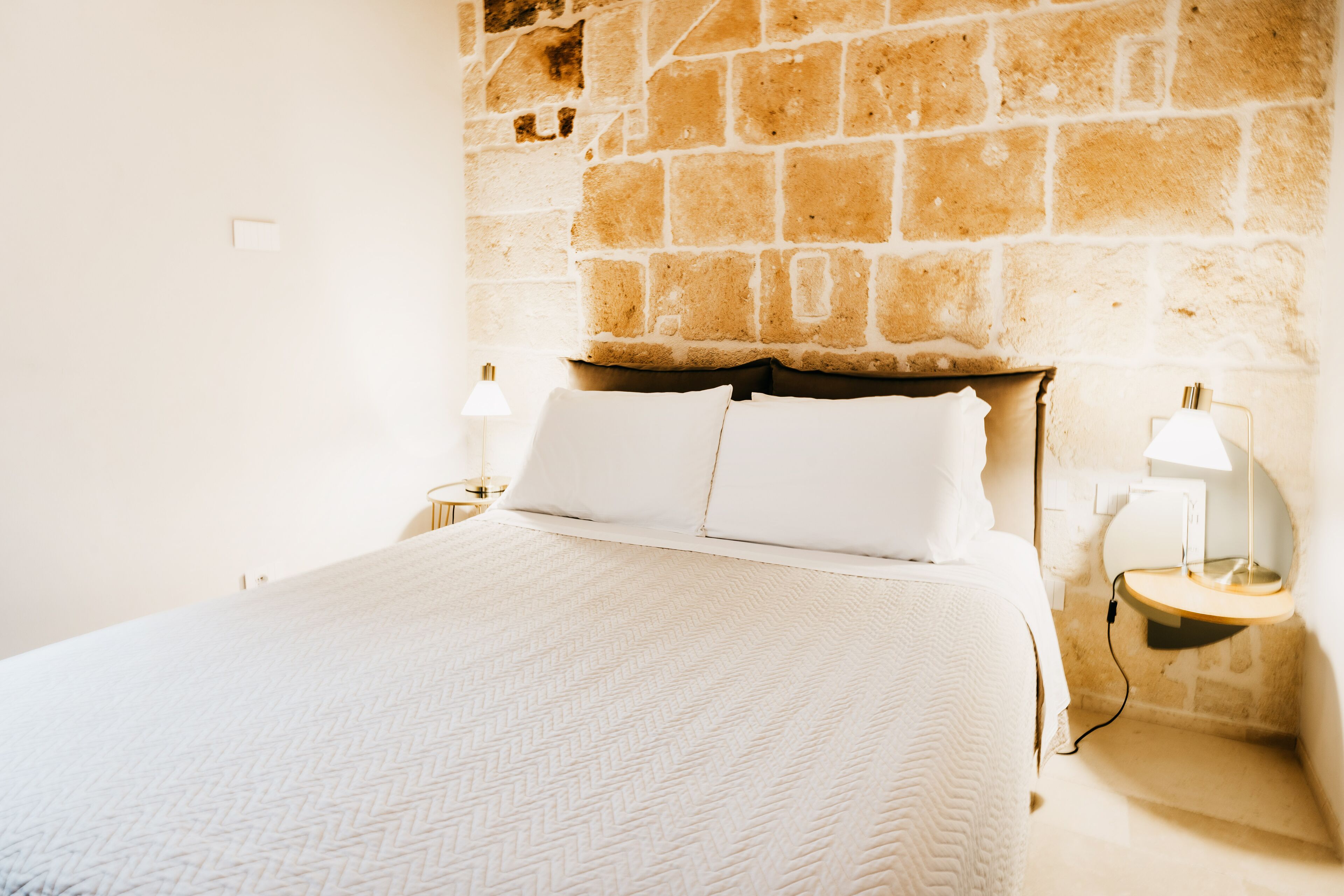 Un lit bien fait, avec du linge de lit blanc, dans une pièce dont un des murs est en pierre.