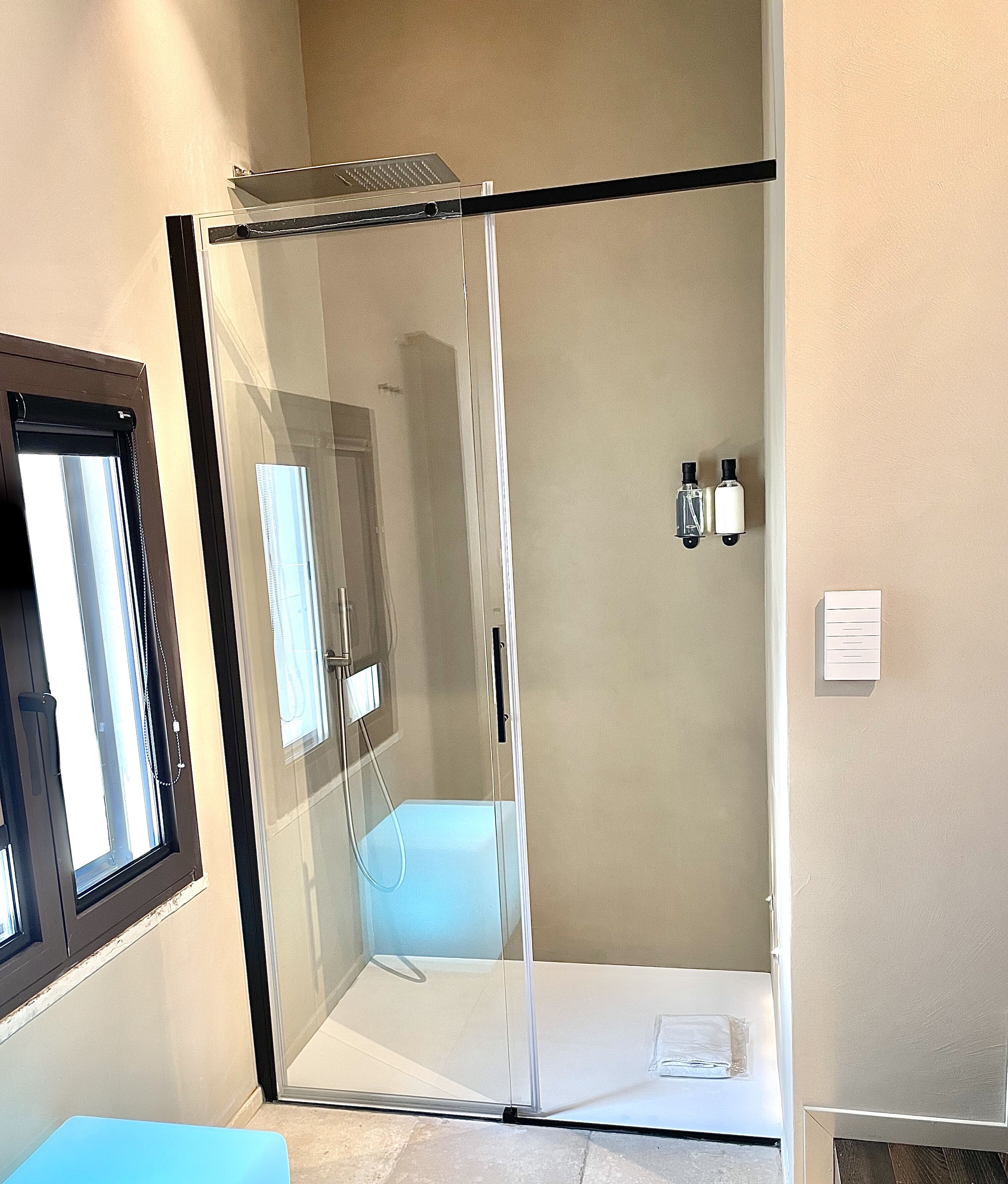 Baño con ducha cerrada por vidrio, un banco, una ventana y un perchero para toallas.