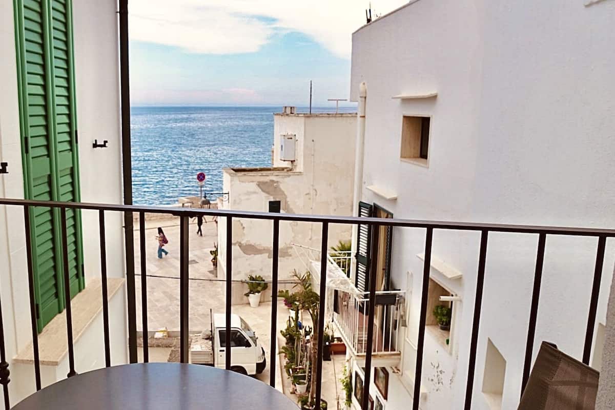 A balcony with a view of the sea, a white building with green shutters, and a parked white van.
