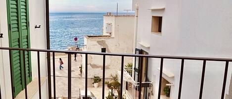 A balcony with a view of the sea, a white building with green shutters, and a parked white van.