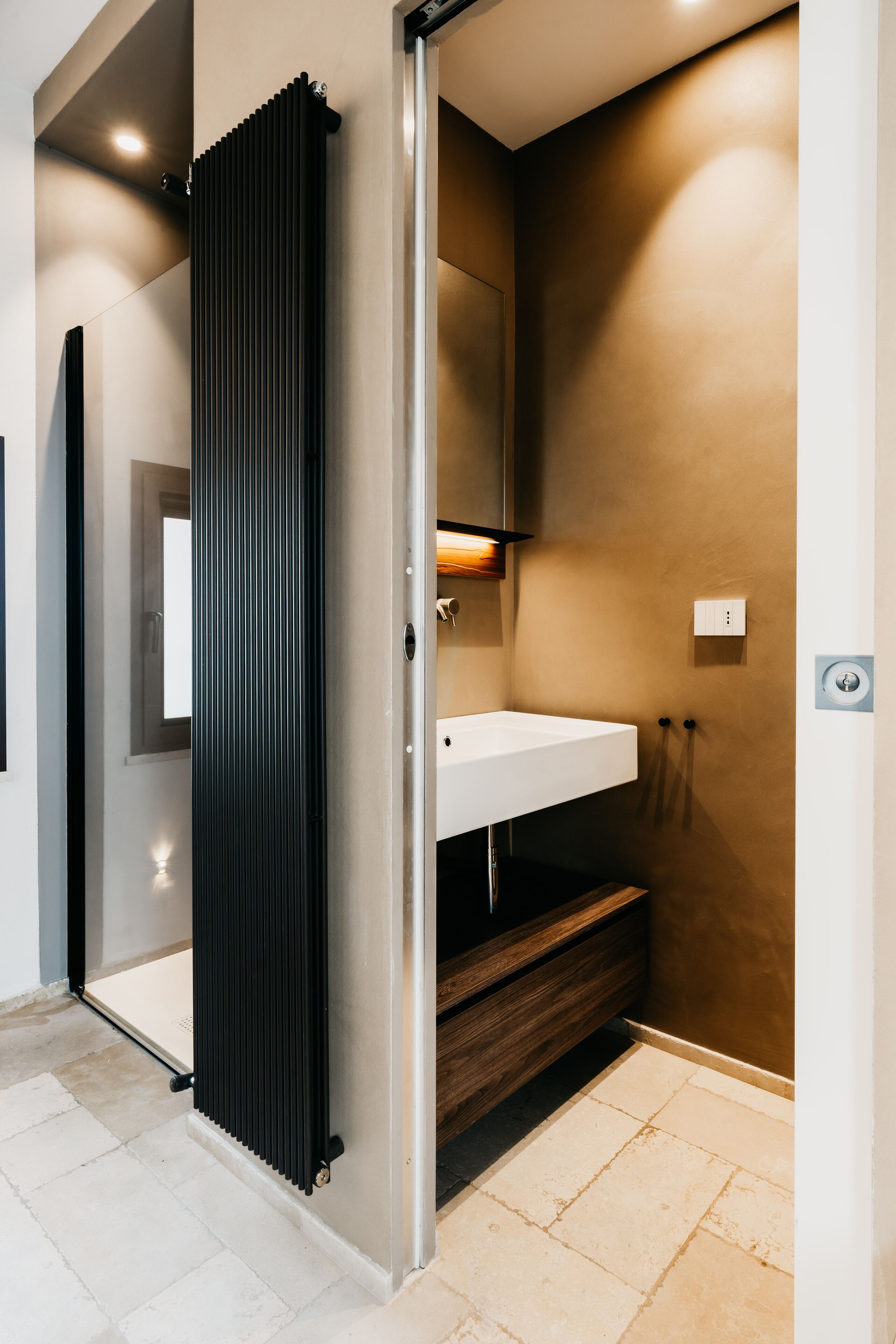Une salle de bain moderne avec un lavabo blanc, un meuble-lavabo en bois, un radiateur noir et un luminaire fixé au mur.