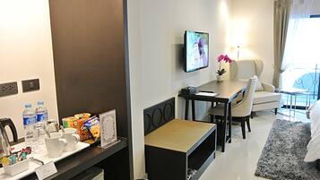 A hotel room with a dining area, a TV mounted on the wall, a small table with a flower vase, a sofa, and a window with curtains.