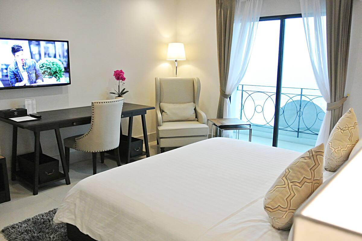 Habitación de hotel con cama, escritorio, silla, televisión y balcón con cortinas.