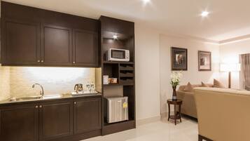 A modern kitchen with dark cabinetry, a stainless steel sink, and a microwave. There is a small refrigerator and a side table with a vase of flowers. A beige sofa is visible in the background.