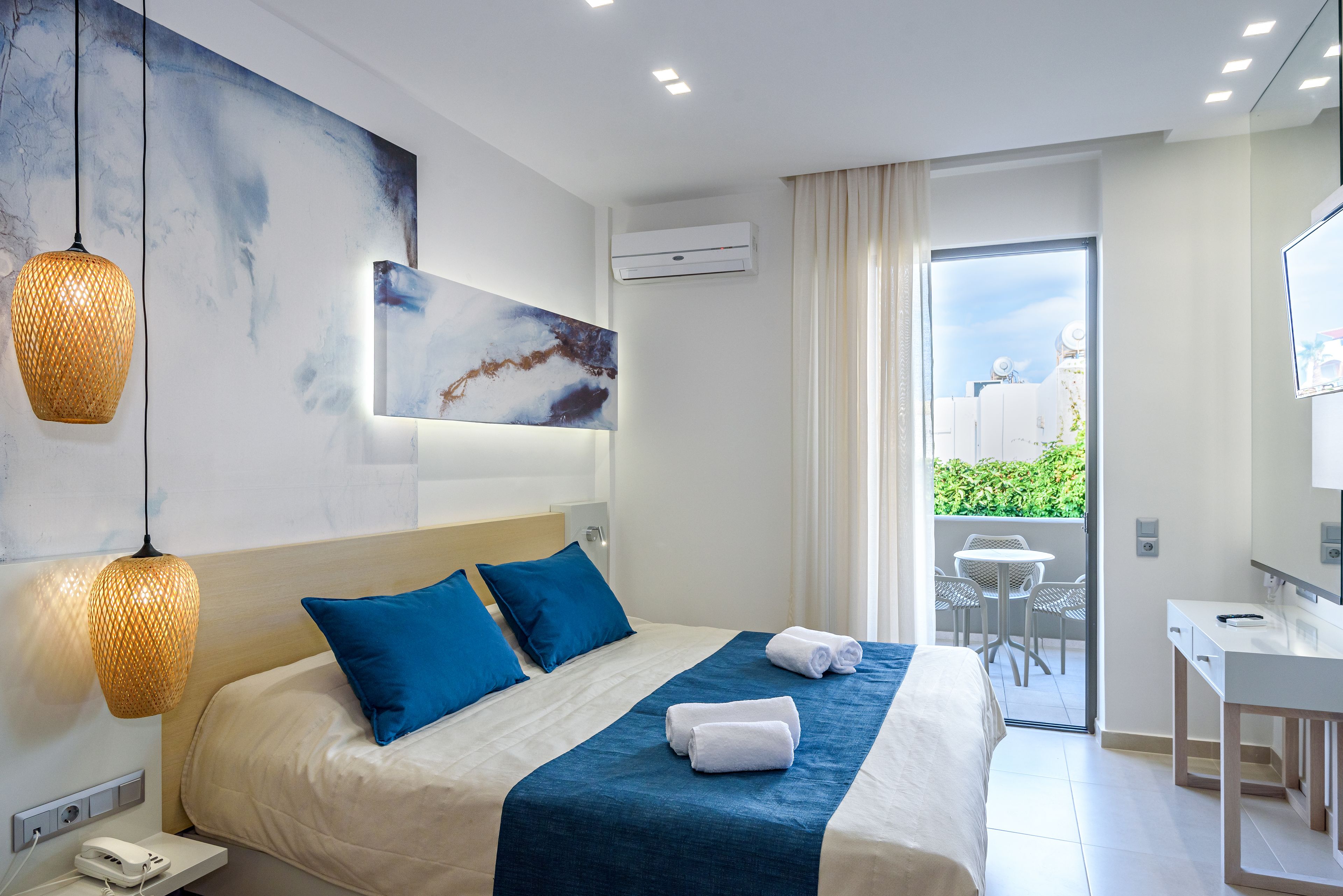 A modern hotel room with a large bed, blue pillows, a desk, and a balcony with outdoor seating.
