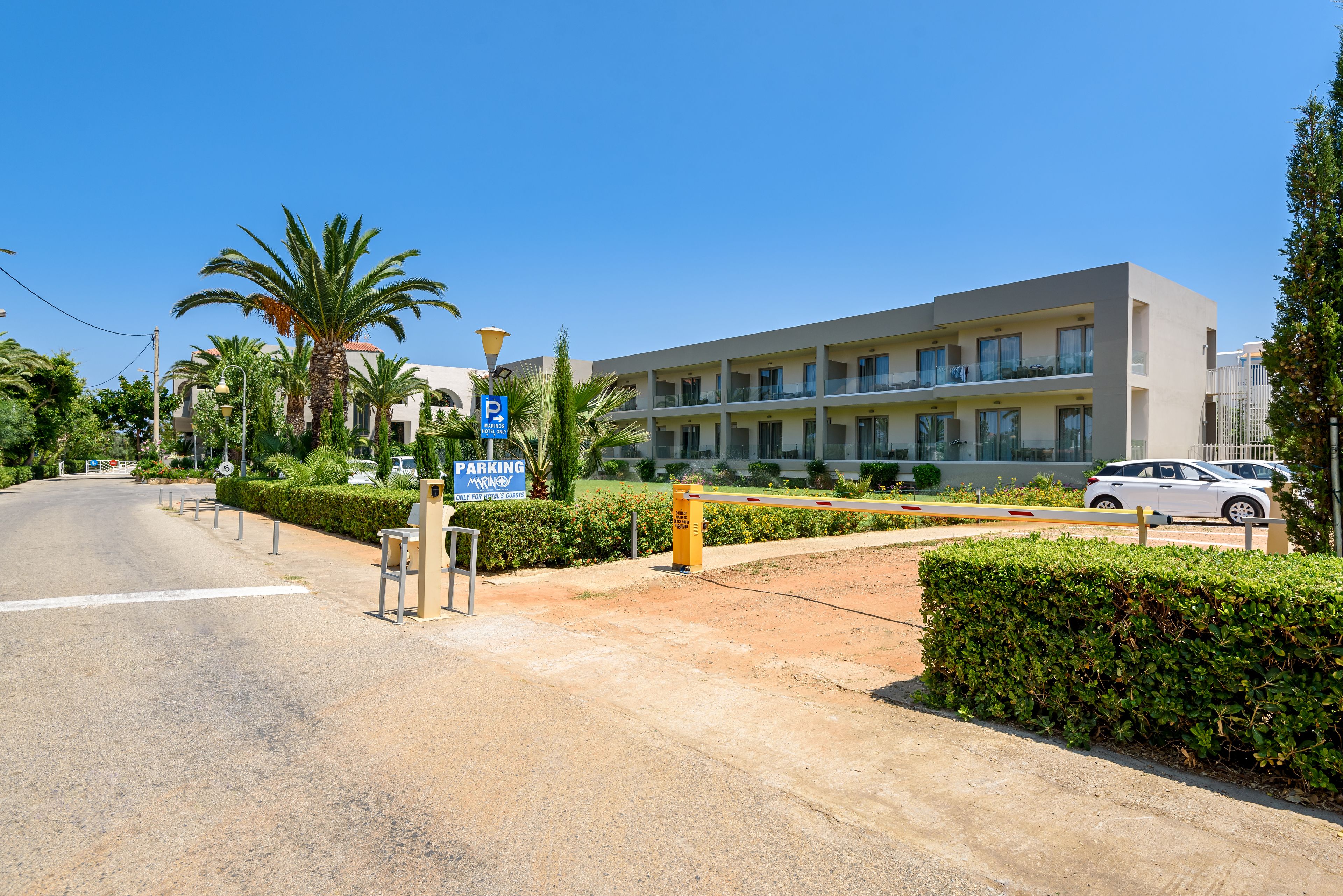 A modern hotel with a parking area, palm trees, and a clear blue sky.