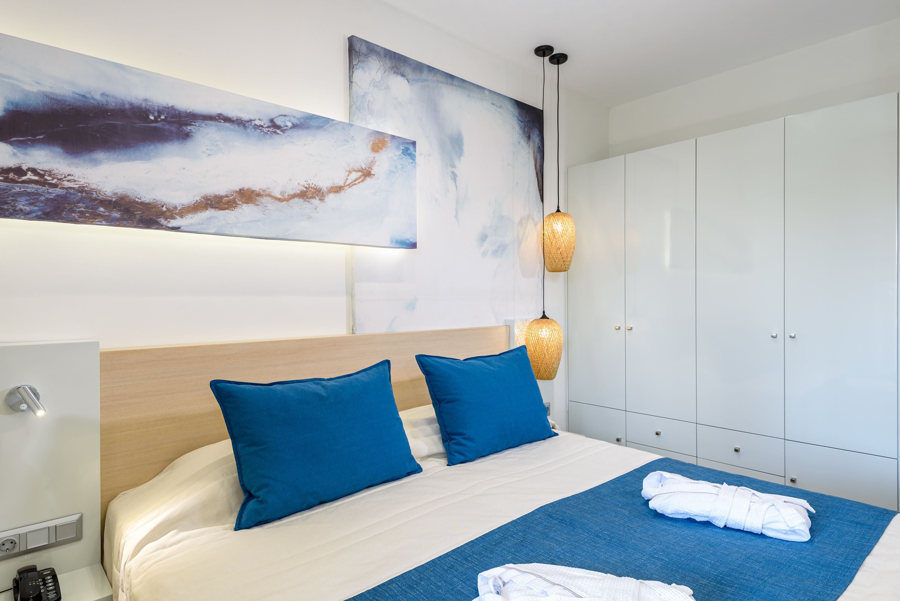 Een moderne hotelkamer met een groot bed, blauwe kussens, een blauw vloerkleed, een witte badjas op het kleed, een groot abstract schilderij, een hanglamp en een kledingkast.