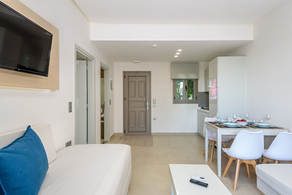 Una habitación de hotel moderna con una cama, un televisor montado en la pared, una zona de comedor con mesa y sillas, y una cocina al fondo.