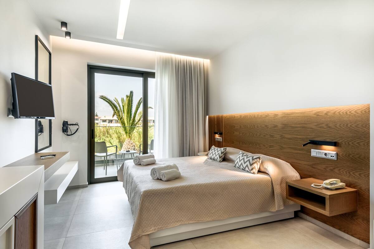 Ein modernes Hotelzimmer mit einem großen Bett, einem an der Wand montierten Fernseher, einem Nachttisch mit Telefon und Blick auf Palmen durch das Fenster.