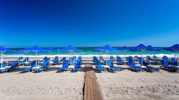 Une plage bordée de rangées de chaises longues et de parasols bleus, une promenade en bois et une vue dégagée sur l’océan.