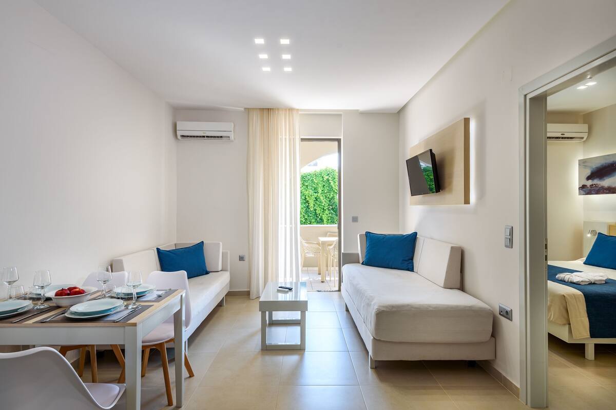 Ein modernes, helles Zimmer mit Essbereich, zwei Sofas, einem kleinen Tisch und einem an der Wand befestigten Fernseher.