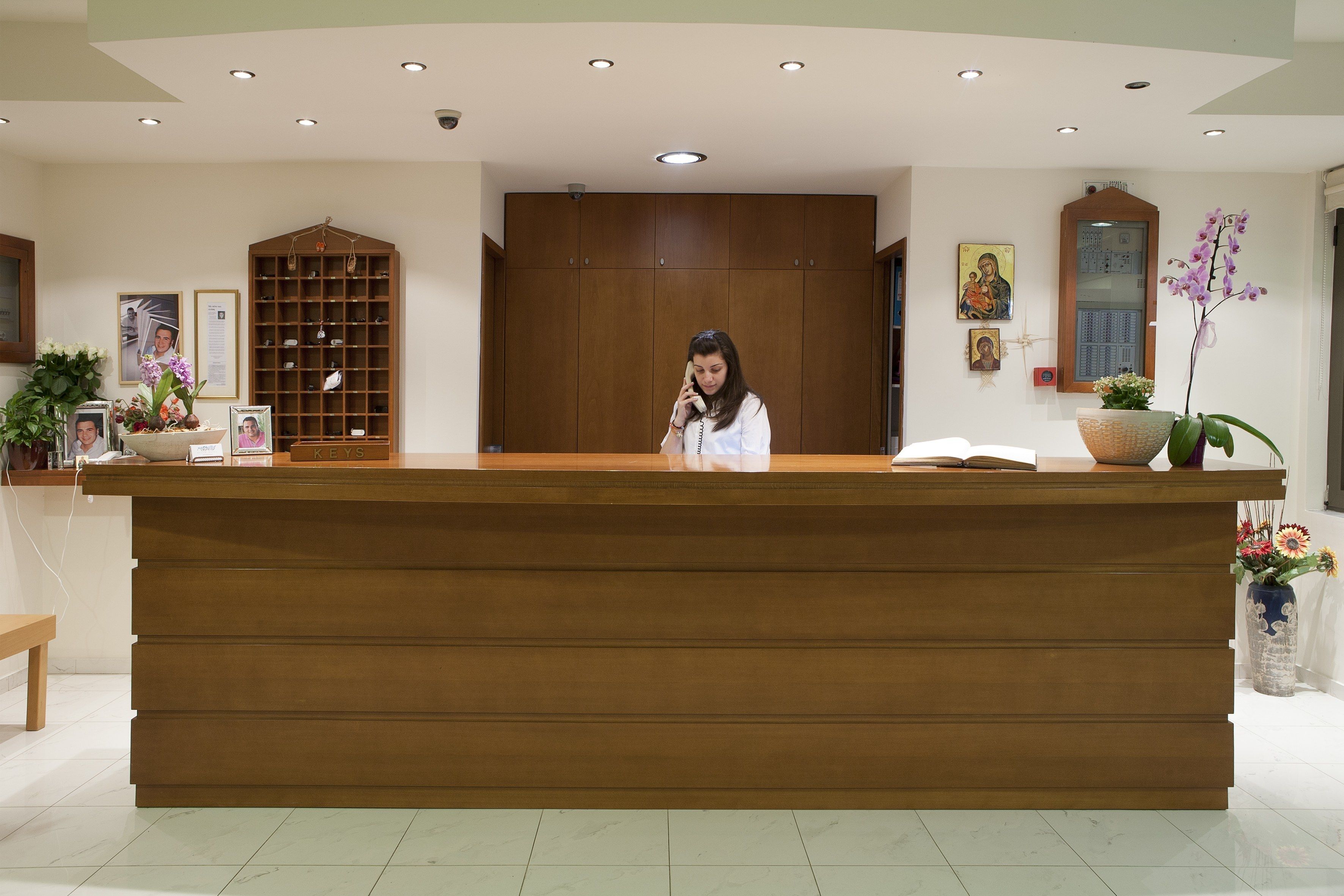 A reception desk with a woman standing behind it, a wooden cabinet, and a wall-mounted shelf with framed photos and a potted orchid.