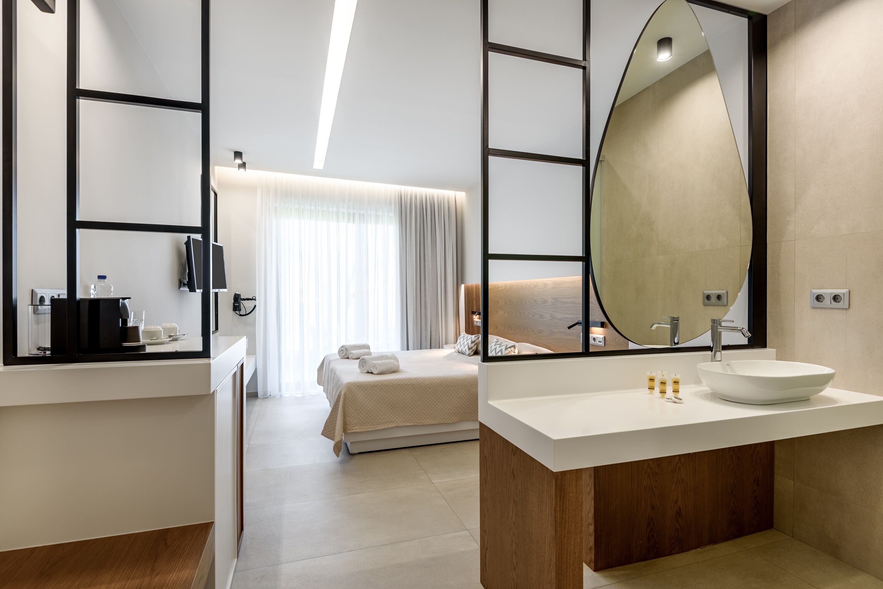 Ein modernes Hotelzimmer mit einem großen Bett, einem Nachttisch mit Kaffeemaschine, einem Spiegel und einem Badezimmer mit großem Waschbecken und Spiegel.
