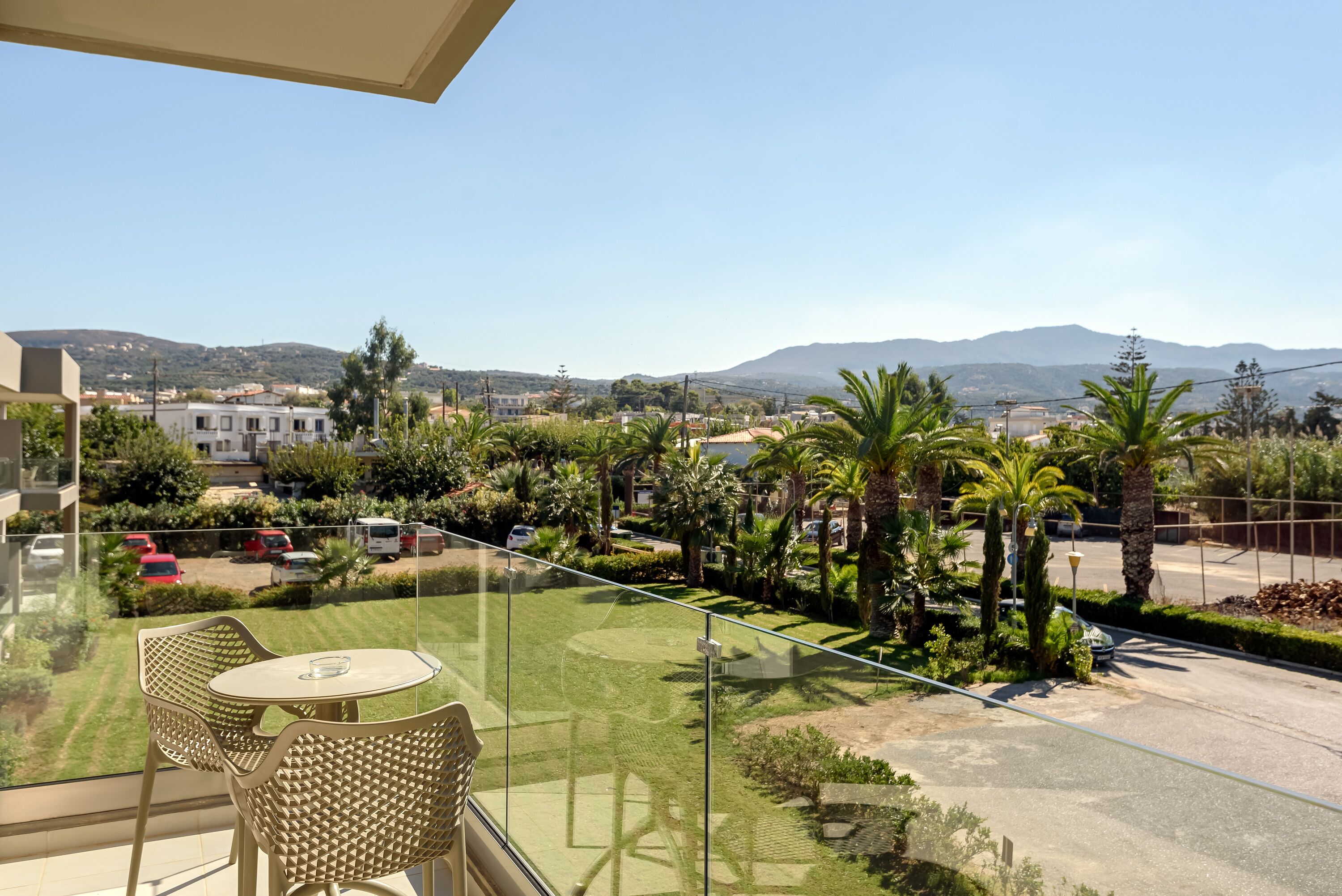 Un balcone con ringhiera in vetro che si affaccia su una zona residenziale con case, palme e cielo sereno.