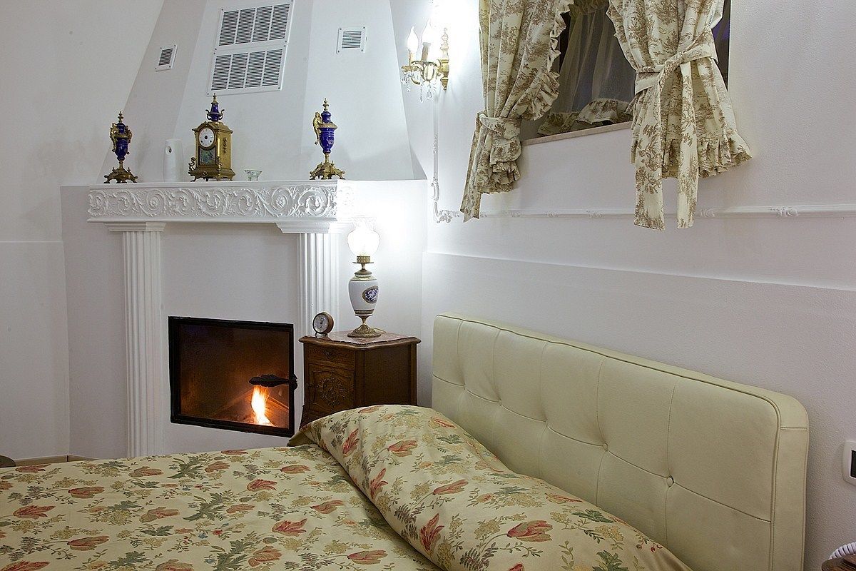 Une chambre avec une cheminée, un lit avec une literie à motifs floraux, une table de chevet avec une lampe et une fenêtre avec des rideaux.