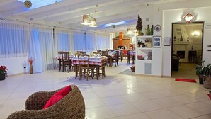 Un amplio comedor con varias mesas y sillas, una silla de mimbre con cojín rojo, y una cocina visible a través de un vano abierto.
