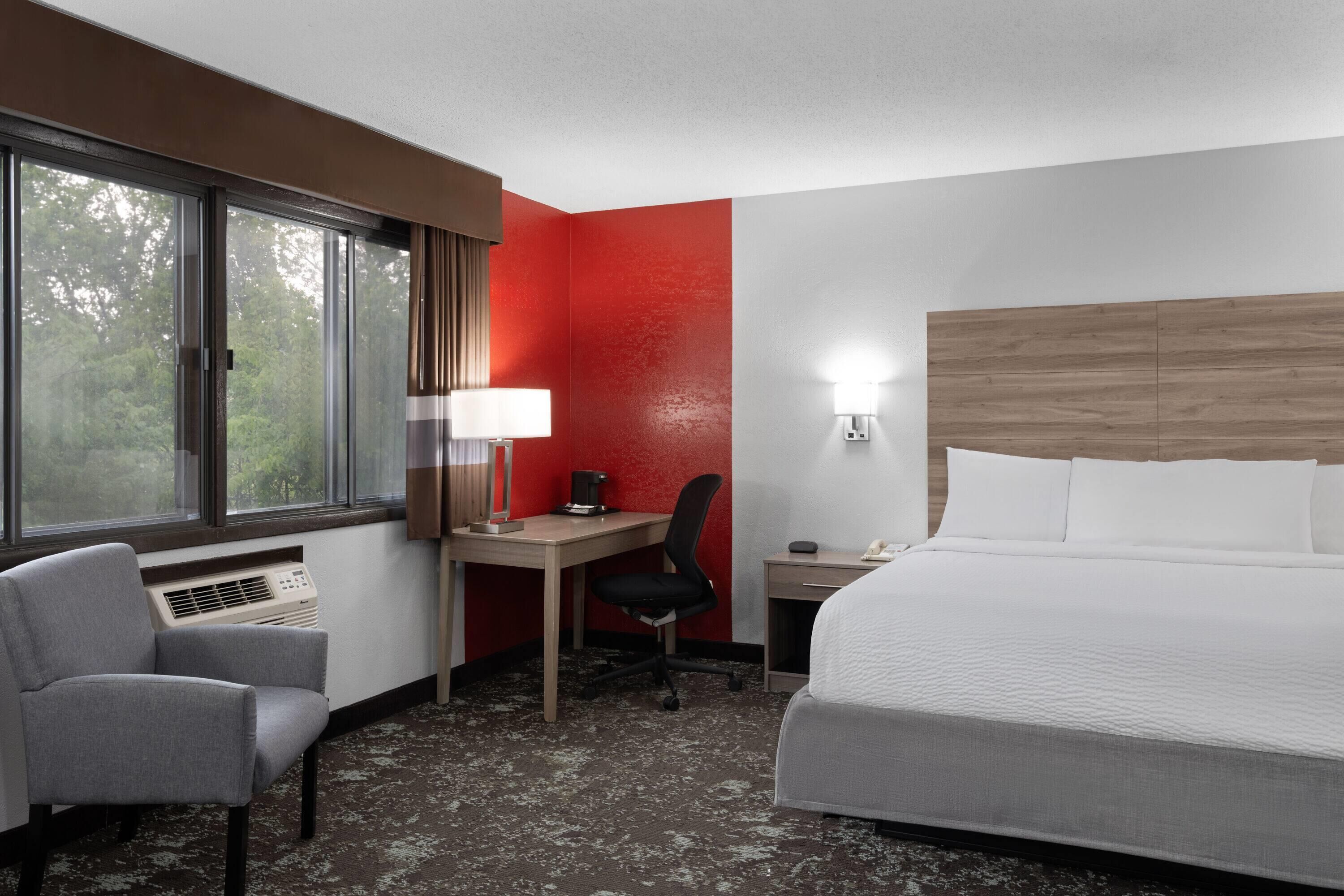 Una habitación de hotel con cama, escritorio, silla, lámpara y ventana con cortinas.