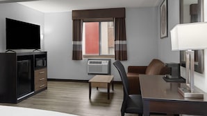 Habitación de hotel compacta con televisión de pantalla plana, minibar, mesa de madera, sofá y escritorio con lámpara.